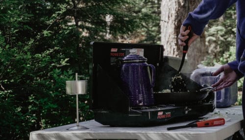 Folding camping tables are perfect for preparing food and cooking outdoors. Person using a folding camping table to cook with a gas camp stove.