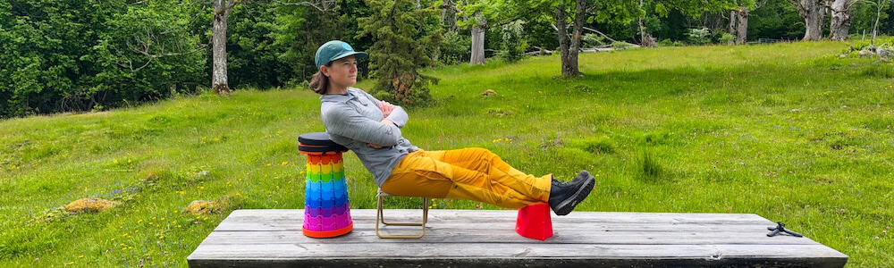 woman sitting on a portable stool using a collapsible chair for a back rest and a pocket chair for a foot rest while hiking and camping